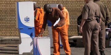 An officer watches on as an inmate inserts his ballot paper into the ballot box at the polling station at Kgoši Mampuru Correctional Facility in Pretoria, South Africa, Wednesday, May 29, 2024. South Africans voted Wednesday at schools, community centers, and in large white tents set up in open fields in an election seen as their country’s most important in 30 years. It could put the young democracy in unknown territory.(AP Photo/Themba Hadebe)